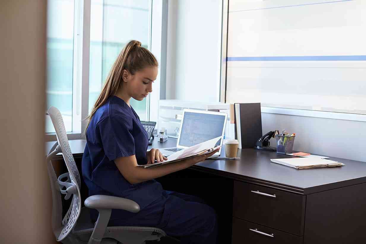 <p>Nurse Wearing Scrubs Working At Desk In Office</p>
