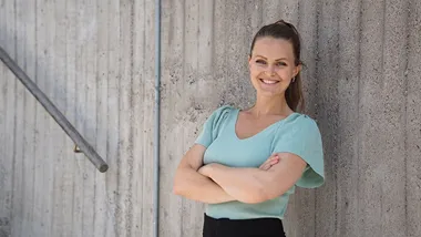 Stina står med armarna i kors lutad mot en grå stenvägg.