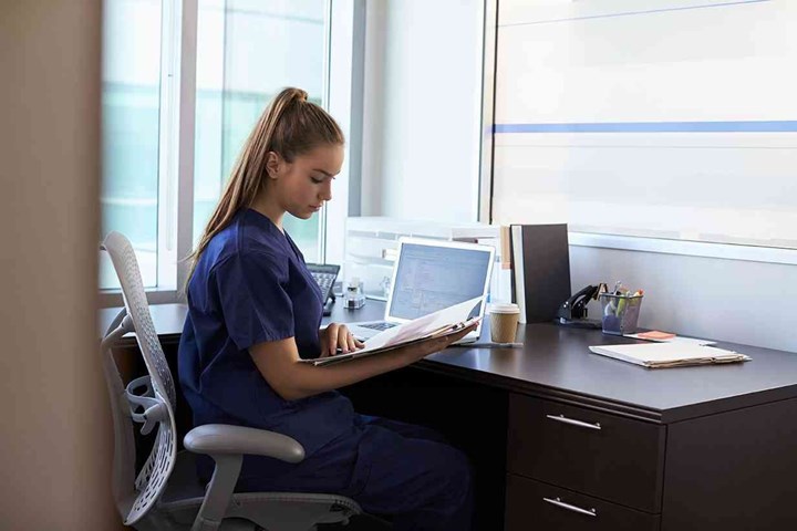 Nurse Wearing Scrubs Working At Desk In Office
