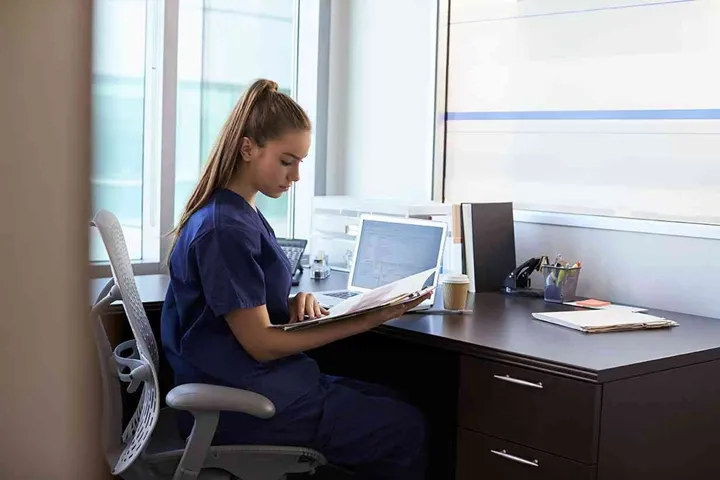 Nurse Wearing Scrubs Working At Desk In Office
