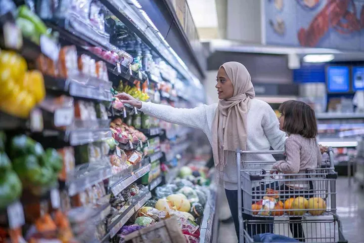 A young Muslim mother and daughter walk the produce aisles of the grocery store together shopping for groceries. They are both dressed casually and the mother is wearing a Hijab. The mother is reaching for products on the shelf while pushing a shopping cart with her daughter seated in it.