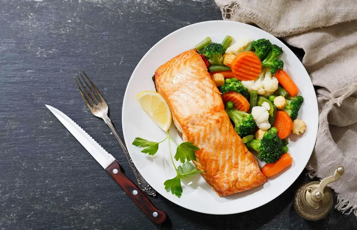 plate of baked salmon steak with vegetables on dark background, top view